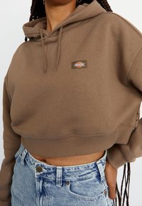 Person som bär en kortärmad brun huvtröja med en liten logotyp och högmidjade ljusblå acid-wash jeans, stående med ena handen på höften.