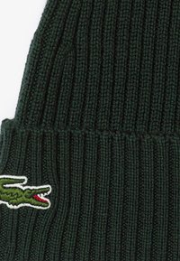 Beanie lavorato a maglia verde scuro con motivo a coste verticali. Presenta un logo di coccodrillo ricamato con bordatura bianca sul lato.