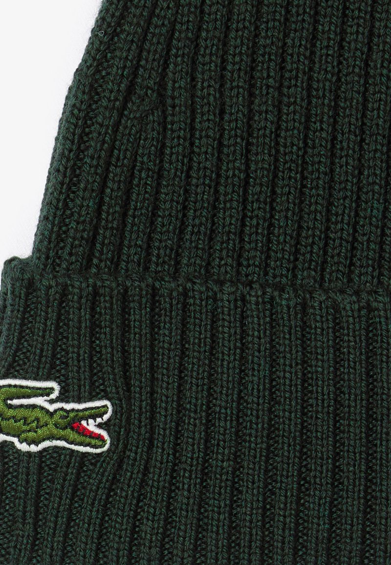 Dunkelgrüne gestrickte Mütze mit vertikalem Rippmuster. An der Seite befindet sich ein weiß umrandetes gesticktes Krokodil-Logo.