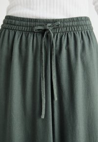 Kalhoty s elastickým pasem v tmavě zelené tkanině s hladkým povrchem, se stahovací šňůrkou pro úpravu pasu.