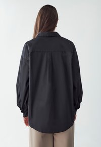 Black oversized shirt with a pointed collar, long balloon sleeves, and buttoned cuffs. Features a rear pleat for added volume.