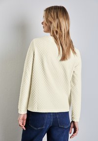 Strukturierter hellgelber Pullover mit langen Ärmeln, rundem Ausschnitt und einem taillierten Saum, kombiniert mit dunkelblauen Jeans.