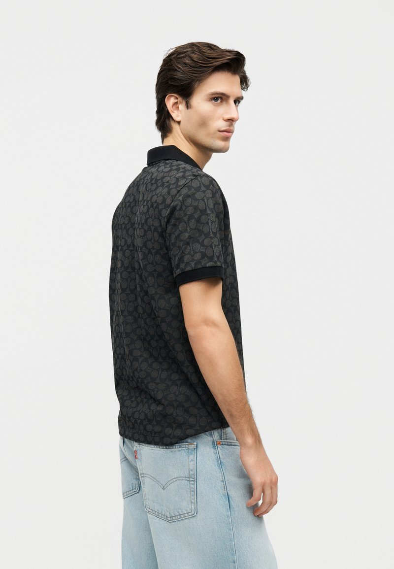 Jeune homme portant un polo noir à motifs et un jean bleu clair, de profil sur un fond uni clair.