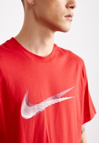 Red cotton t-shirt featuring a large, stylized white Nike logo across the front. The shirt has a classic crew neck design.