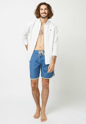 Camicia leggera bianca a righe verticali sopra pantaloni da bagno blu. I pantaloni presentano un laccio e un bordino a contrasto. Nessuna scarpa visibile.