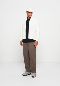 Giacca bianca con zip sopra una maglietta nera, pantaloni a righe marroni e scarpe grigie. Un cappellino chiaro completa l'outfit.
