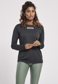 Langærmet sort atletisk skjorte med et hvidt logo på brystet, parret med grønne leggings. Glat tekstur, tætsiddende design.