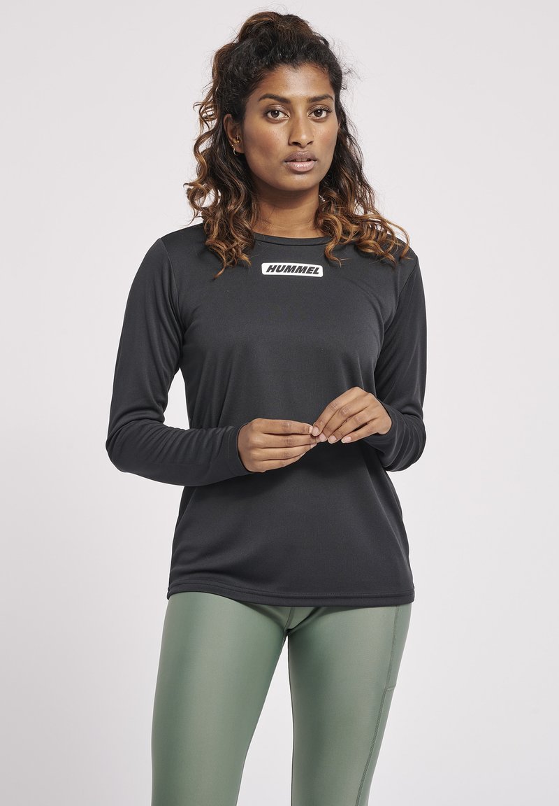 Langærmet sort atletisk skjorte med et hvidt logo på brystet, parret med grønne leggings. Glat tekstur, tætsiddende design.