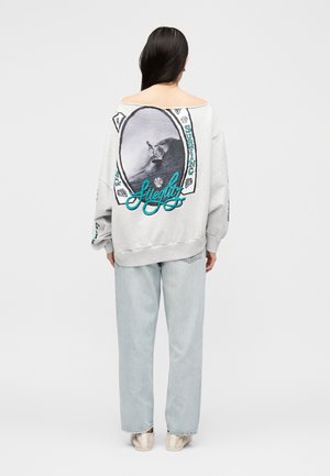 Femme aux longs cheveux foncés portant un sweat-shirt gris clair avec un grand graphique de surf et un texte turquoise au dos, un jean bleu clair et des chaussures beiges.