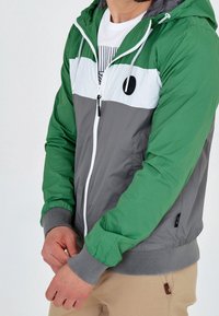 Kurtka przeciwwiatrowa z zielonymi i szarymi panelami, białym paskiem, zamek błyskawiczny, kaptur ze sznurkiem oraz elastycznymi mankietami. Teksturowany, lekki materiał.