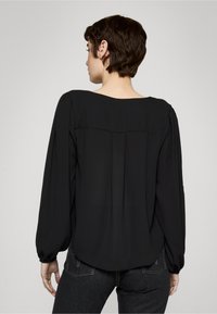 Blusa negra de gasa con mangas largas abullonadas, escote amplio y detalle plisado en la espalda, combinada con jeans de mezclilla oscura.