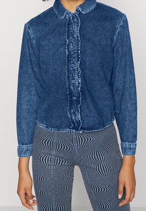 Camicia in denim con fronte arricciato, maniche lunghe e colletto arrotondato. Accompagnata da pantaloni a fantasia neri e blu.