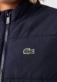 Giacca imbottita blu navy con colletto alto, caratterizzata da un logo di coccodrillo ricamato in verde e bianco e una cerniera argentata. Texture morbida.