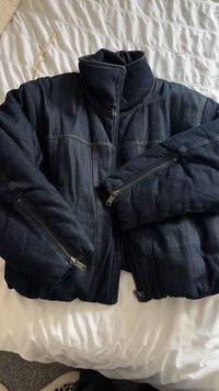 Tmavě modrá džínová bunda s vysokým límcem, kapsami na zip a vycpávanými rukávy. Vyznačuje se viditelným šitím a strukturovaným designem.