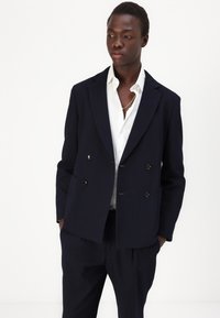 Blazer doppiopetto blu navy con rever a punte e due tasche frontali. Abbinato a una camicia bianca, con una texture liscia e linee pulite.