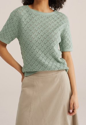 Femme portant un haut tricoté vert clair à manches courtes avec un motif géométrique et une jupe beige taille haute, main sur la hanche.