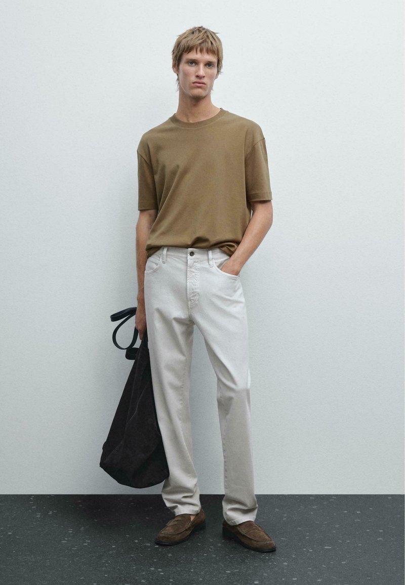 Maglietta in cotone marrone, pantaloni in denim bianchi, scarpe in suede marroni e una borsa nera. L'uomo è in piedi contro uno sfondo chiaro e texturizzato.