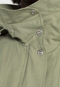 Jasnozielona kurtka z wysokim kołnierzem, z metalowymi napami i teksturowanym materiałem. Prezentuje skomplikowane detale w szwie.