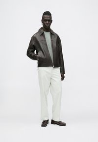 Brūns ādas žakete ar asu apkakli, zem tā pelēks džemperis, gaišas krāsas bikses un tumši apavi. Gluda tekstūra ar rāvējslēdzēja aizdari.