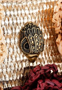 Gevlochten gouden en beige clutch met een zwarte en gouden decoratieve sluiting die lijkt op een vogel. Opvallend door de gestructureerde materialen en gevlochten accenten.