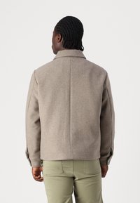 Bruine wolmixjas met een kraag, details op de manchetten en een relaxte pasvorm. Gepaard met olijfgroene broeken, getoond vanaf de achterkant.