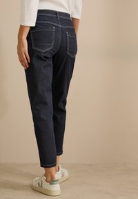 Jeans en denim bleu foncé avec des coutures blanches, design à jambes effilées et deux poches arrière. Portés avec des baskets de couleur claire.