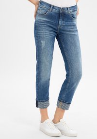 Jean en denim bleu avec une coupe décontractée et des poignets retroussés, dévoilant une doublure intérieure à motifs. Comprend des détails de couture et cinq poches.