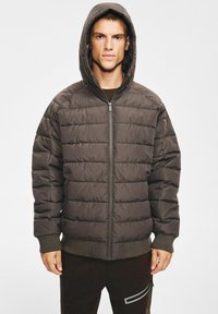 Ciemnobrązowa pikowana kurtka puchowa z zamkiem błyskawicznym, dużym kapturem, ściągaczami na rękawach i pikowaną fakturą. Model nosi ją na ciemnych spodniach.