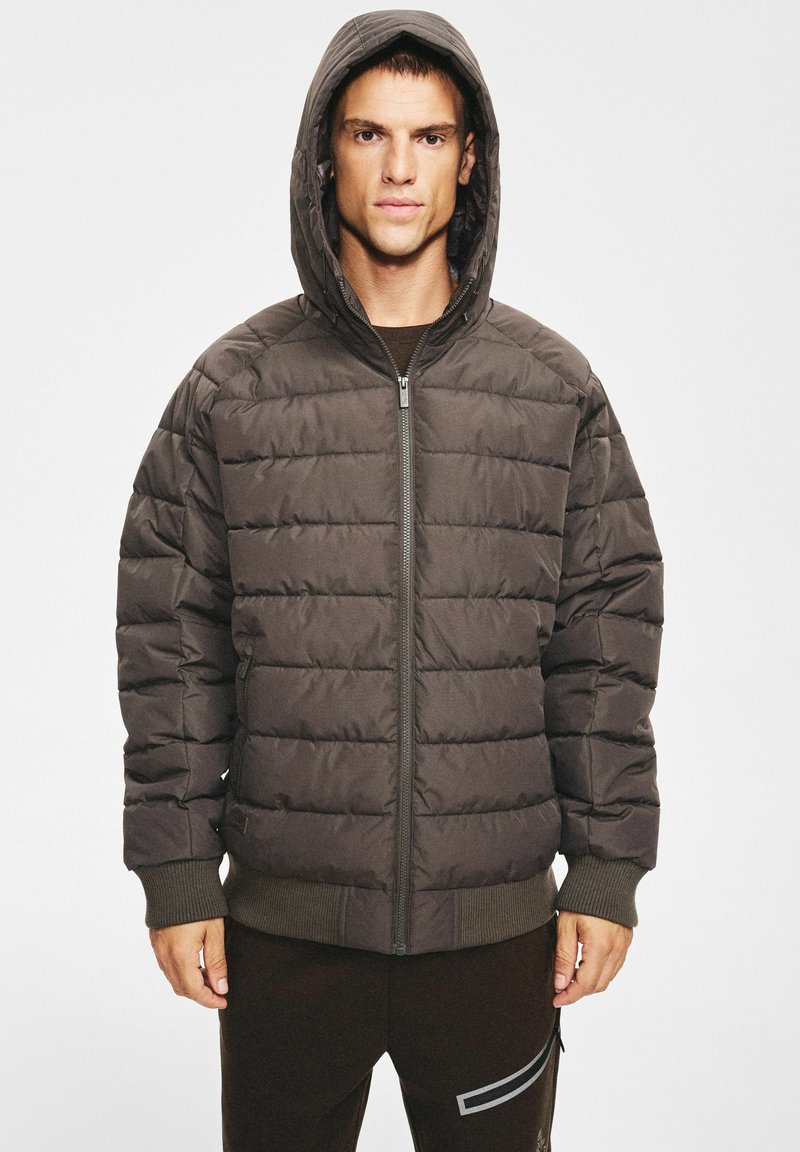 Ciemnobrązowa pikowana kurtka puchowa z zamkiem błyskawicznym, dużym kapturem, ściągaczami na rękawach i pikowaną fakturą. Model nosi ją na ciemnych spodniach.