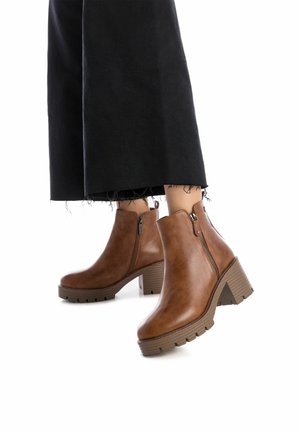 Botas de cuero marrón hasta el tobillo con un tacón блок y suela texturizada; detalles de cremallera lateral y un acabado suave. Combinadas con pantalones negros recortados.