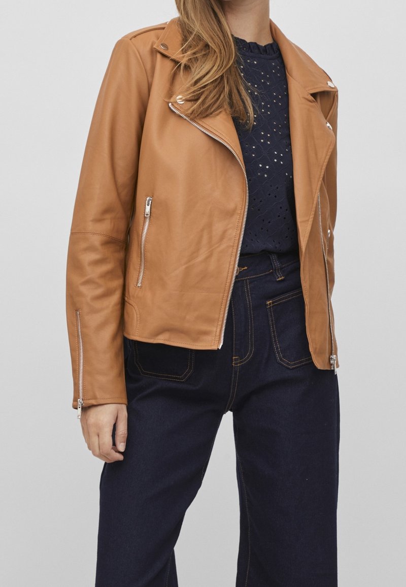 Brązowa skórzana kurtka motocyklowa z srebrnymi zamkami błyskawicznymi, z dopasowanym krojem, skróconą długością i subtelnymi szczegółami w stębnowaniu. Noszona na granatowej bluzce.