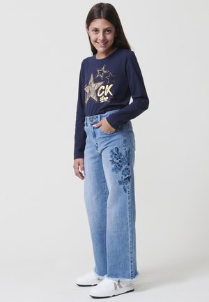 Chica sonriendo, vistiendo una camiseta de manga larga azul marino con estrellas doradas, jeans azul claro con bordados florales azules, zapatillas blancas, con la mano en el bolsillo.