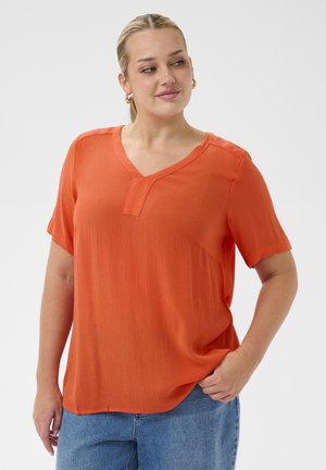 Žena s blond vlasmi v copu, ktorá nosí zlaté kruhové náušnice, oranžovú blúzku s krátkym rukávom a modré džínsy, stojí s jednou rukou v vrecku.