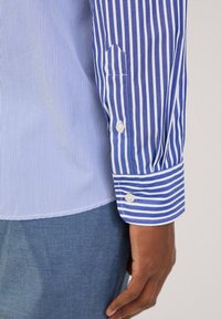 Camisa a rayas con rayas verticales en azul claro y blanco, con puños de botón y un detalle de puño puntiagudo. Textura de tela de algodón suave.