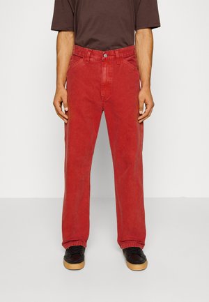 Osoba ubrana w czerwone, luźne spodnie, ciemnobrązową koszulkę z krótkim rękawem oraz czarne sneakersy z gumowymi podeszwami, stojąca na szarej podłodze.