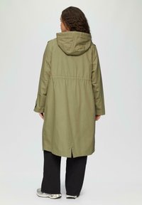 Een vrouw met krullend haar, gekleed in een olijfgroene lange jas met capuchon, zwarte wijde broek en witte sneakers, staat met de rug naar de camera tegen een effen achtergrond.