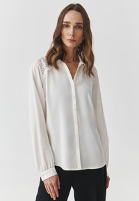 Blusa blanca con botones en la parte delantera, mangas largas y acentos de encaje en los hombros. Textura suave y corte relajado.