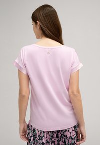 Roze top met korte mouwen en een ronde halslijn, met opgerolde mouwen. De achterkant heeft een klein ankerdetail bij de halslijn. Gladde stofstructuur.