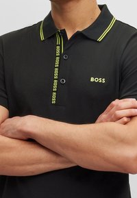 Hombre vestido con una camiseta polo negra con el logo "BOSS" en color verde neón en el pecho y los botones, con los brazos cruzados sobre el torso.