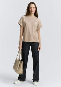 T-shirt en coton beige avec un col rond et des manches courtes, associé à un pantalon ample en denim foncé et un sac à main beige clair. Baskets blanches.