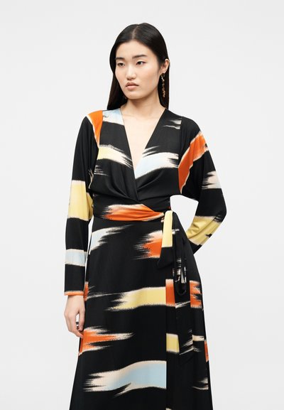 Γυναίκα με μακριά σκούρα μαλλιά που φοράει μαύρο φόρεμα τύπου wrap με αφηρημένα μοτίβα πινελιές σε πορτοκαλί, κίτρινο και μπλε, στέκεται μπροστά σε απλό φόντο.