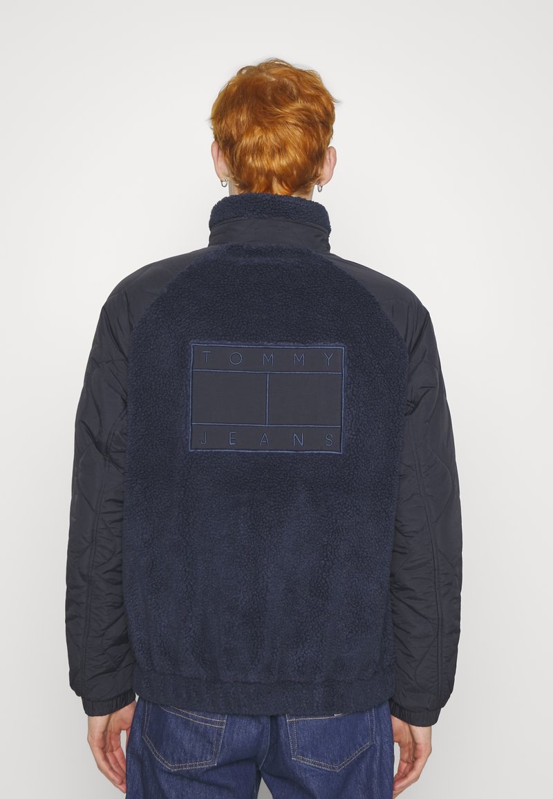 Granatowa kurtka z polarem z tyłu, pikowanymi rękawami, wysokim kołnierzem i haftowanym logo "TOMMY JEANS" na plecach. Miękka tekstura i luźny krój.