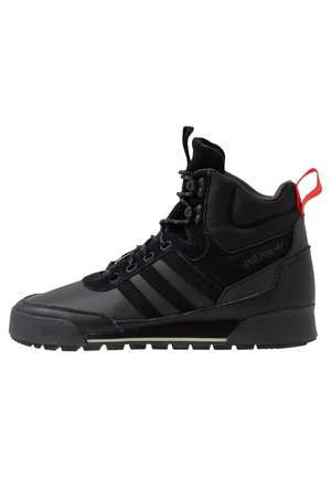 Czarny sneaker za kostkę z paskami Adidas, z przodu na sznurowadła, o wytrzymałej podeszwie, z wyściełanym kołnierzem wokół kostki oraz czerwoną pętelką na pięcie.