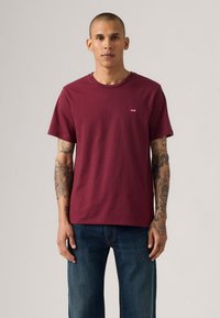 T-shirt bordeaux in cotone con colletto a girocollo, maniche corte e un piccolo logo rosso sul lato sinistro del petto. Indossato con jeans in denim blu.