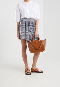 Bolso de ante marrón con dos asas superiores, falda mini con patrón geométrico en azul oscuro y blanco, y sandalias marrones con acentos metálicos.