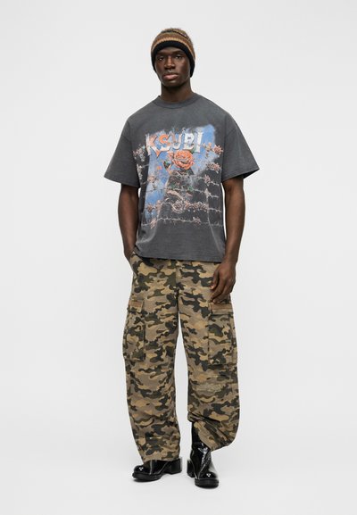 Γκρι γραφικό μπλουζάκι με σχέδιο τριαντάφυλλο, σετ με φαρδιά παντελόνια καμουφλάζ cargo και μαύρες δερμάτινες μπότες. Το μοντέλο φοράει πλεκτό καπέλο.