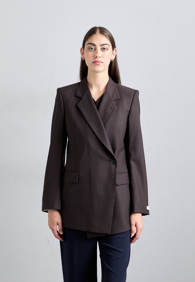 Brown double-breasted blazer featuring sharp lapels, structured shoulders, and two front pockets, paired with dark trousers.