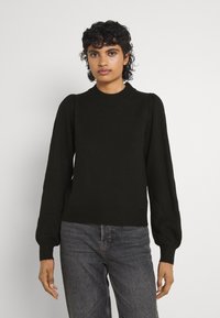 Maglione nero lavorato a maglia con scollatura rotonda, maniche a sbuffo e orlo a coste. Abbinato a jeans in denim scuro. Sfondo neutro.
