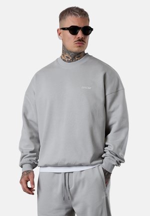 Hellgraues Sweatshirt mit geripptem Rundhalsausschnitt und Bündchen, versehen mit einem kleinen Logo. Über einer weißen Schicht getragen, kombiniert mit passenden Jogginghosen.