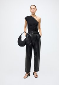 Schwarzes One-Shoulder-Top kombiniert mit locker sitzender schwarzer Lederhose, die eine taillierte Passform mit Gürtel aufweist. Dazu eine schwarze, geschwungene Handtasche und Riemchensandalen.